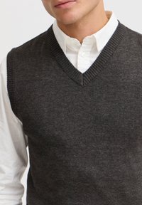 Gilet en tricot gris foncé avec encolure en V et bords côtelés, porté par-dessus une chemise blanche à col, mettant en valeur une texture lisse et une coupe ajustée.