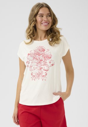T-shirt crème à manches courtes avec un motif floral rouge. Tissu doux avec une coupe décontractée. Associé à un pantalon rouge, porté de manière décontractée.