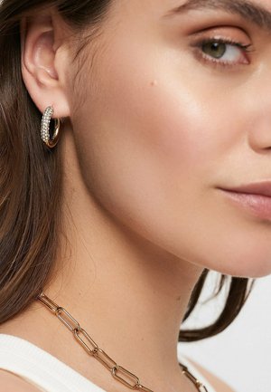 Primo piano del volto di una donna che mostra orecchini a cerchio dorati con cristalli e una collana a catena spessa in oro su pelle chiara.