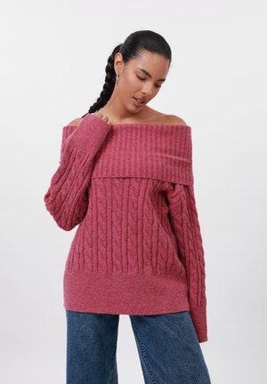 Off-the-shoulder, geribbelde trui in een diepe roze kleur met kabelbrei textuur. Lange mouwen en een losse pasvorm, gecombineerd met blauwe jeans.