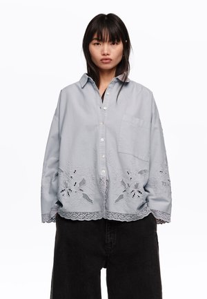 Joven con cabello negro y liso que lleva una camisa azul claro de botones de gran tamaño con bordados florales y pantalones negros de pierna ancha.