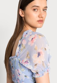 Blusa floral azul claro com mangas bufantes transparentes, detalhe de corpete franzido e padrões florais em cores pastel. Tecido suave e leve.