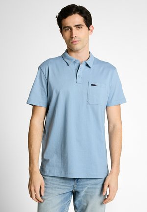Hombre con polo de manga corta azul claro con bolsillo, de pie contra fondo blanco liso, mirando hacia adelante.