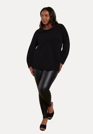 Musta pitkähihainen paita, jossa on rento malli, yhdistettynä kiiltävän mustiin leggings-housuihin ja avokauluksisiin mustiin korkokenkiin, seisoo vaaleaa taustaa vasten.