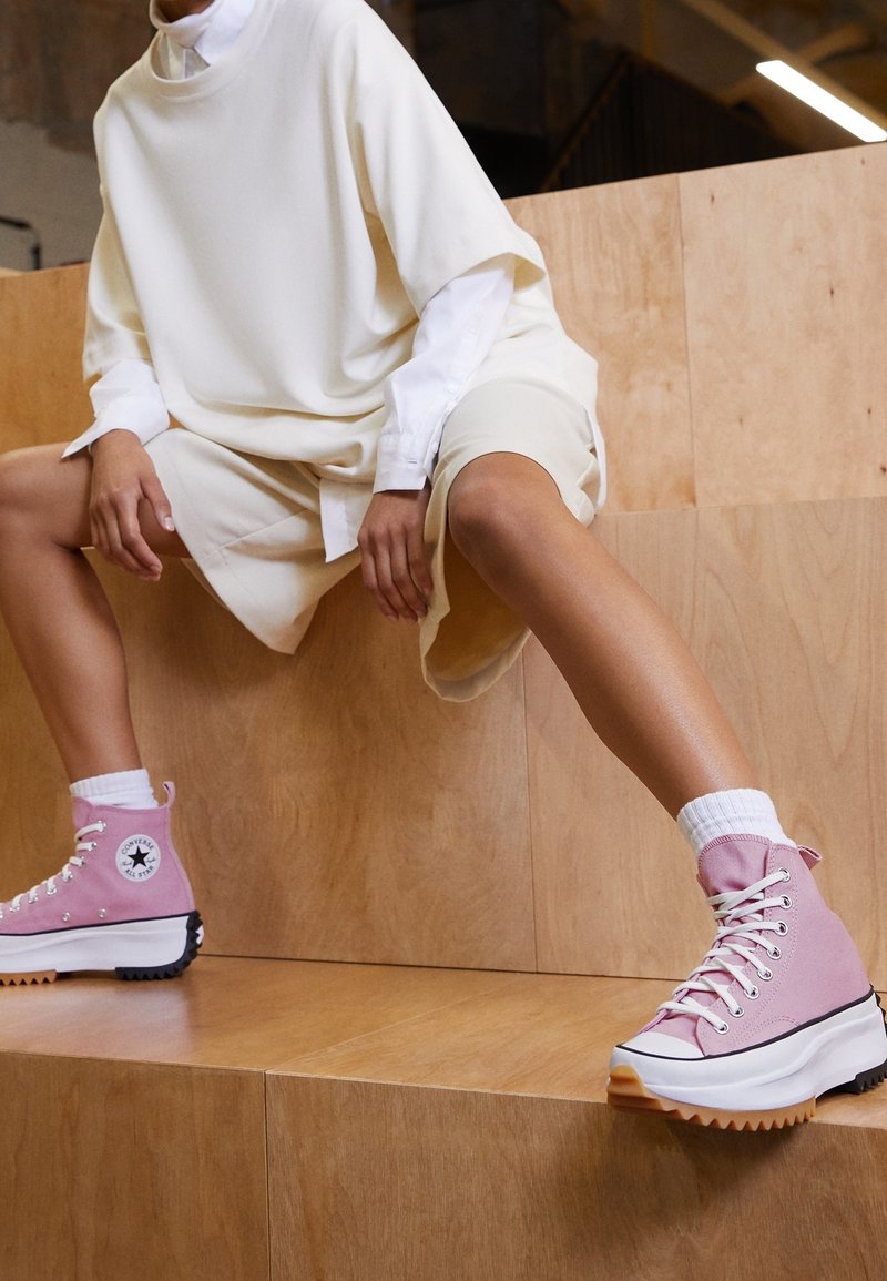 Pink high-top sneakers with a chunky white sole, black outsole, and white laces. The model wears a beige oversized top and shorts.