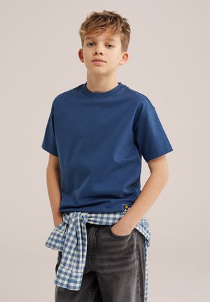 Chłopiec z krótkimi blond włosami, ubrany w niebieski t-shirt i czarne jeansy, z niebiesko-białą koszulą w kratę zawiązaną wokół talii.