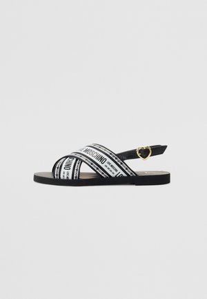 Flad sort sandal med hvide krydsede remme trykt med teksten "Love Moschino" og en guldfarvet hjerteformet spænde på ankelremmen.
