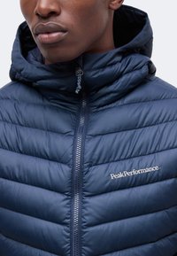 Homme portant une veste matelassée bleu marine avec une capuche et un logo "Peak Performance" visible sur la poitrine.