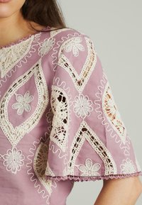 Rosa Bluse mit komplexer cremefarbener Spitzenstickerei, Blumenmustern, gewellten Kanten und ausgeschnittenem Detail an den Ärmeln.