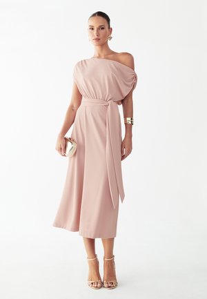 Naine kannab tuhmunud roosa üheõlalist midi-pikka kleiti, millel on vöökinnis, kuldseid kõrvarõngaid, käevõru, väikesekotti ja kõrgete kontsadega sandaale valgel taustal.