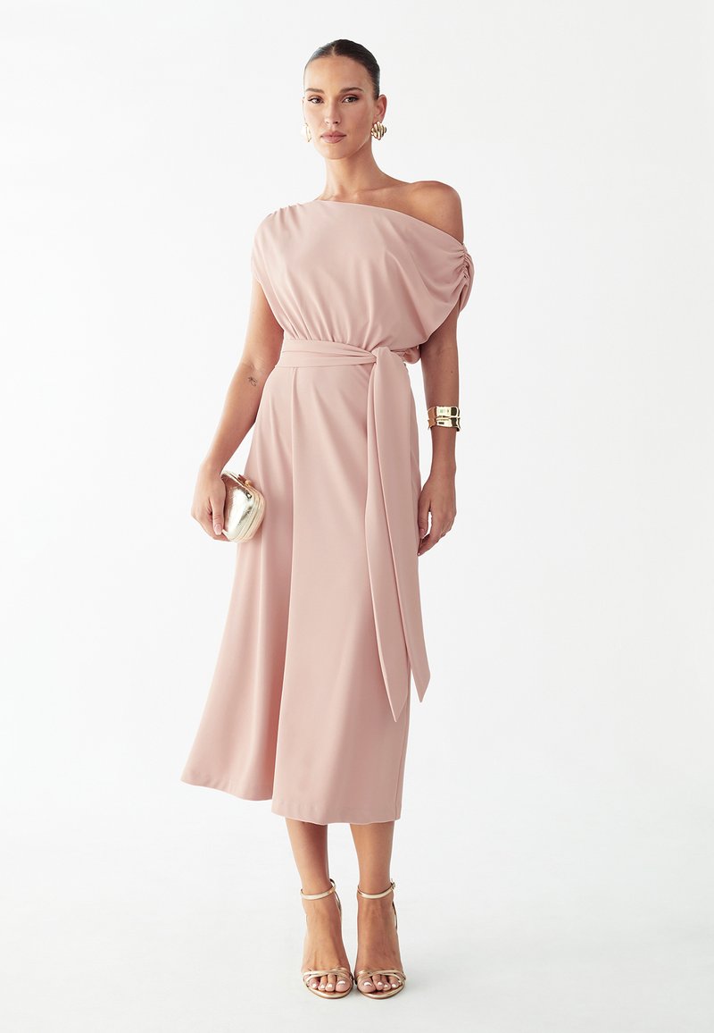 Žena v ružovkastoružových midi šatách na jedno rameno so zaväzovaním v páse, zlatými náušnicami, náramkom, kabelkou a sandálmi na vysokom opätku na bielom pozadí.