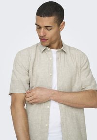 Camisa de lino ligera de manga corta en color beige claro con cuello abotonado, que presenta una textura suave y un patrón de tejido sutil. Lleva una camiseta blanca debajo.