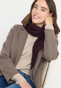 Écharpe bordeaux avec des accents texturés portée sur un pull beige et un manteau gris. Caractéristiques : coupe décontractée et poignets de manches côtelés.