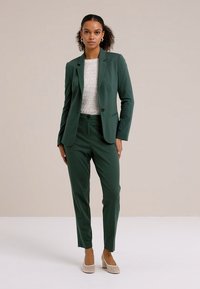 Tamsiai žalia pritaikyta kostiuminė apranga su vieno mygtuko švarku ir siaurėjančiomis kelnėmis, dėvima ant šviesaus tekstūruoto megztinio, derinama su smėlio spalvos lakuotais bateliais.