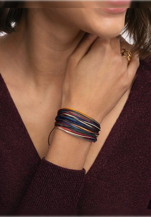 Donna che indossa un braccialetto a fili multicolore a strati e un anello d'oro sulla mano destra, vestita con un maglione bordeaux a scollo a V.