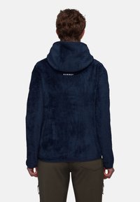 Mörkblå fleecejacka med texturerad yta och huva. Har varumärket "Mammut" placerat på baksidan av halsringningen.