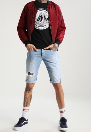 Giubbotto bomber bordeaux con polsini a costine, t-shirt nera con grafica, pantaloni corti in denim strappati di colore blu chiaro e scarpe da ginnastica blu navy con suola bianca.