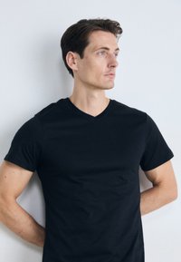 Jeune homme aux cheveux bruns courts portant un T-shirt noir à col en V, se tenant les mains derrière le dos, regardant vers la droite devant un mur blanc uni.