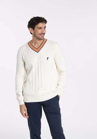 Pull en maille torsadée crème avec décolleté en V, comportant un col rayé orange et noir. Petit logo brodé sur la poitrine gauche.