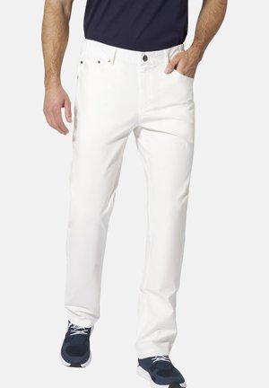 Mann trägt weiße gerade geschnittene Hose, marineblaue Sneakers und ein dunkles Oberteil, steht mit einer Hand in der Tasche vor einfarbigem Hintergrund.
