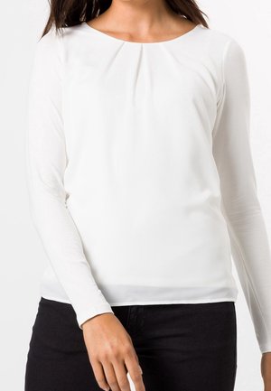 Femme portant un chemisier blanc ajusté à manches longues avec un détail plissé au niveau de l'encolure ronde et un pantalon noir.