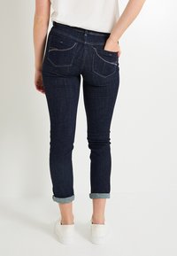 Pantalones vaqueros skinny de denim azul oscuro con dobladillos acampanados, dos bolsillos traseros y detalles de costura sutiles. Llevados con zapatos blancos sin cordones.