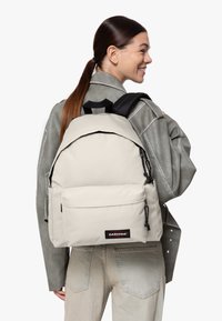 Béžový batoh Eastpak zaobleného tvaru, s kapsou na zip na přední straně a nastavitelnými černými popruhy, nošený přes šedou bundu.