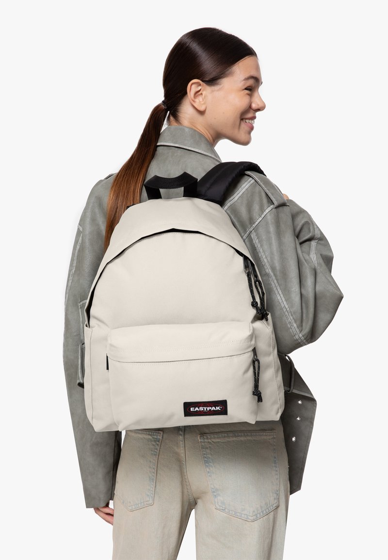 Béžový batoh Eastpak zaobleného tvaru, s kapsou na zip na přední straně a nastavitelnými černými popruhy, nošený přes šedou bundu.