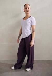 Haut ajusté violet clair avec des boutons, associé à un pantalon large violet foncé et des chaussures blanches. Mis en valeur sur un fond neutre.