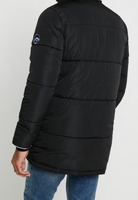 Giubbotto impermeabile nero con pannelli a cuscino orizzontali, colletto alto e logo blu di una montagna sulla manica sinistra. Materiale testurizzato e resistente all'acqua.