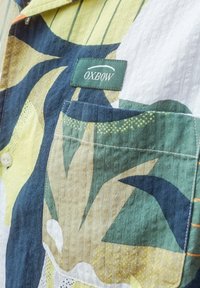 Chemise à manches courtes avec poche plaquée, fabriquée en tissu texturé présentant un motif floral abstrait multicolore dans des tons de vert, jaune et bleu marine.