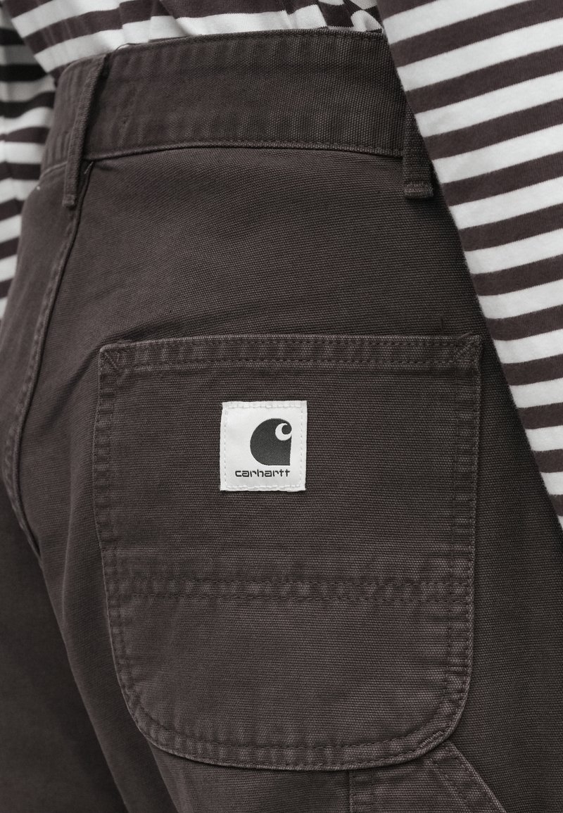 Primer plano del bolsillo de unos pantalones Carhartt marrón oscuro con etiqueta de la marca, llevados con una camiseta a rayas negras y blancas.