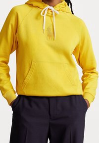 Gul hoodie med framficka, vita snoddar och broderad detalj. Ribbad mudd vid ärmar och nederkant, mjukt tyg med en avslappnad passform.