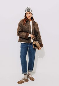 Femme portant une veste marron, un pull blanc, un jean bleu retroussé, un bonnet rayé, des baskets à motif léopard, et tenant un chapeau marron contre un mur uni.