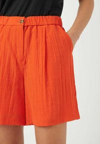 Oranssi laskostetut shortsit, joissa on joustava vyötärö, edessä nappi ja sivutaskut, valmistettu teksturoidusta, kevyestä kankaasta.