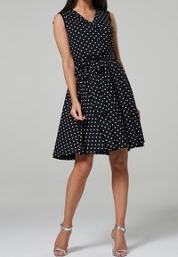 Femme portant une robe noire sans manches longueur genou à pois blancs, taille nouée et sandales argentées à talons hauts, debout devant un fond gris.