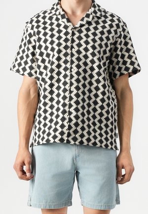 Mand iført sort- og hvidternede kortærmede skjorte med knapper og lyseblå denimshorts mod hvid baggrund.