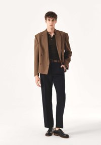Rjava dvorezenska jakna čez črno polo majico, v kombinaciji z mornarsko modrimi hlačami in črnimi usnjenimi čevlji. Gladek material, krojena postava.
