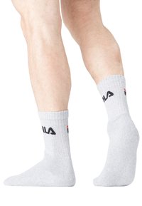Chaussettes de sport grises avec texture côtelée, arborant un logo "FILA" en noir et des accents rouges. Visibles sur un pied avec un ton de peau clair.