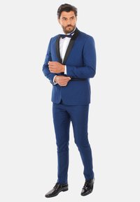 Smoking blu con rever in satin nero, camicia bianca e papillon nero. Design aderente, tessuto liscio e scarpe nere lucide.