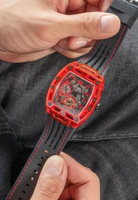 Caja de reloj rectangular transparente roja con correa de goma negra y roja. Presenta una esfera con múltiples diales, números en negrita y acentos texturizados.