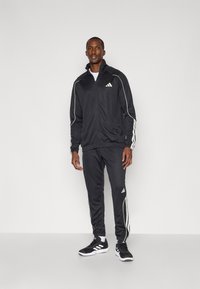 Melna sportiska treniņtērpa komplekts, kas izgatavots no gludas auduma, ar baltām svītrām uz piedurknēm un biksēm, ar jaku ar rāvējslēdzēju un Adidas logotipa detaļām.