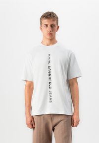 Jaunas vyras dėvi baltą marškinėlį su vertikaliais juodais užrašais "KARL LAGERFELD JEANS" ir šviesiai rudomis kelnėmis, stovėdamas prieš baltą foną.