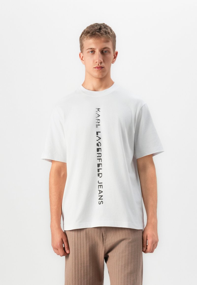 Ungdommelig mann i hvit T-skjorte med vertikal svart tekst "KARL LAGERFELD JEANS" og lysebrune bukser, stående mot en hvit bakgrunn.