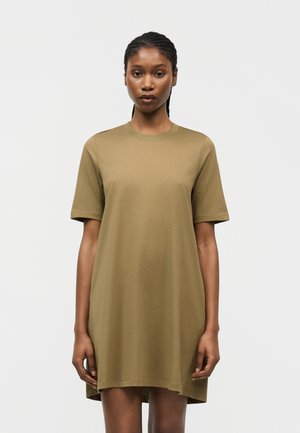 Kobieta z warkoczami, ubrana w luźną, krótką, oliwkowozieloną sukienkę-tunikę, stojąca na jasnym, jednolitym tle.