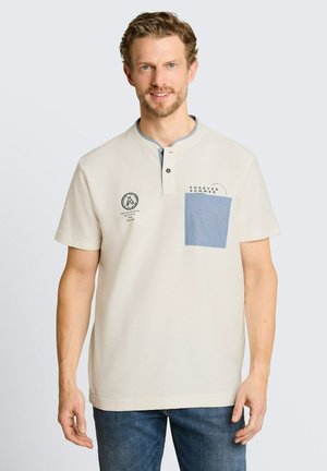 Mann mit kurzen lockigen Haaren, der ein beiges Kurzarmhemd mit einer blauen Brusttasche und bedrucktem Text trägt, stehend vor einem einfarbigen Hintergrund.