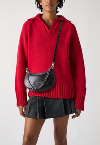 Suéter de punto rojo con cuello, combinado con una falda negra con pliegues y un pequeño bolso negro con cadena y logotipo.