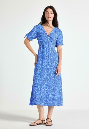 Donna che indossa un vestito midi blu con stampa floreale bianca, maniche corte con laccetti e sandali piatti marroni, in piedi davanti a uno sfondo chiaro e uniforme.