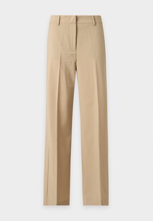 Person trägt beige Anzughose mit weitem Bein und Blazer, schwarzes Hemd, hält eine schwarze Handtasche und trägt rote Schuhe, steht vor neutralem Hintergrund.