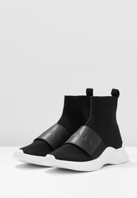 Černé tenisky ve stylu ponožek s bílými gumovými podrážkami. Mají černý pás s embosovaným logem napříč střední částí. Bezšvíková pletená textura.